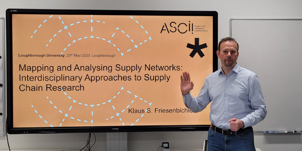 A person standing beside a large screen displaying a presentation slide. The slide reads: 'ASCII, Loughborough University, 20th May 2025, Loughborough. Mapping and Analysing Supply Networks: Interdisciplinary Approaches to Supply Chain Research. Klaus S. Friesenbichler.